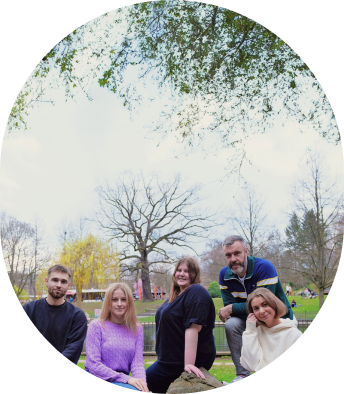 Fünf Menschen sitzen und stehen entspannt in einem Park mit Bäumen und blauem Himmel.