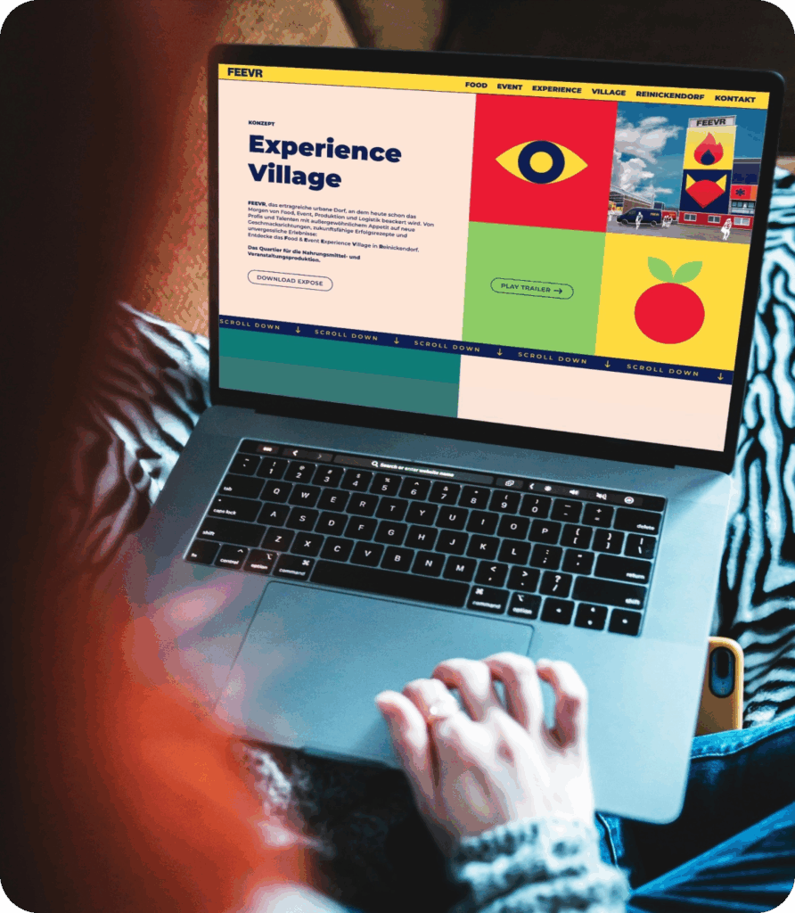 Person sitzt mit Laptop auf dem Schoß und betrachtet eine Webseite mit dem Titel 'Experience Village' in deutscher Sprache, die bunte farbige Blöcke und Illustrationen zeigt, darunter ein Auge, eine rote Frucht und ein Gebäude mit dem Logo 'FEEVR'