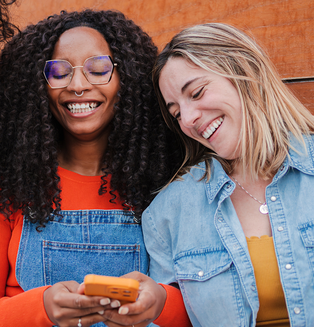 Zwei lachende Frauen, eine mit lockigen schwarzen Haaren und Brille trägt ein oranges Shirt und Jeans-Latzhose, die andere mit blonden Haaren trägt ein gelbes Top und eine blaue Jeansjacke, beide schauen auf ein orangefarbenes Handy, vor einer Holzwand