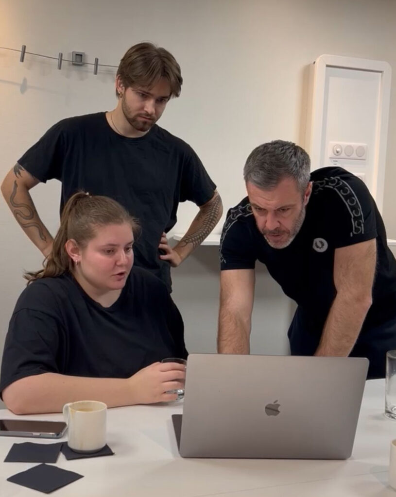 Drei Personen in schwarzer Kleidung blicken konzentriert auf einen Laptop vor ihnen auf einem weißen Tisch, eine Frau sitzt und hält ein Glas Wasser, zwei Männer stehen daneben mit Tattoos auf den Armen, im Hintergrund eine weiße Wand mit einer Steckdosenleiste.