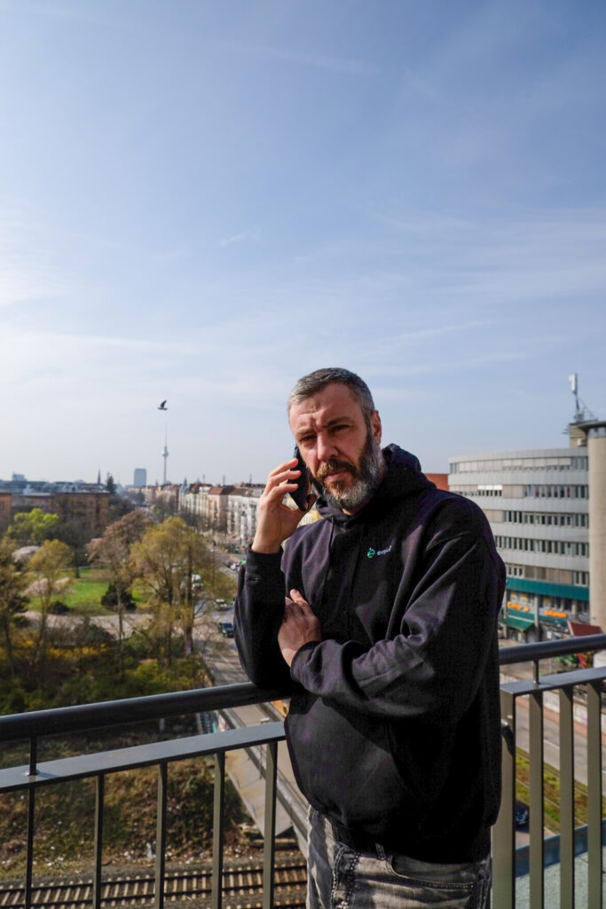 Mann mit Bart und schwarzem Hoodie, der auf einem Balkon steht und in einem sonnigen Stadtbild mit Bäumen und Gebäuden telefoniert.