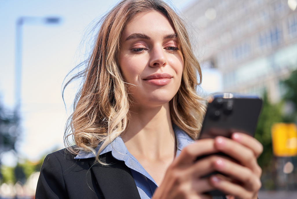 Junge Frau mit blonden Haaren in Businesskleidung blickt lächelnd auf ihr Smartphone im Freien.